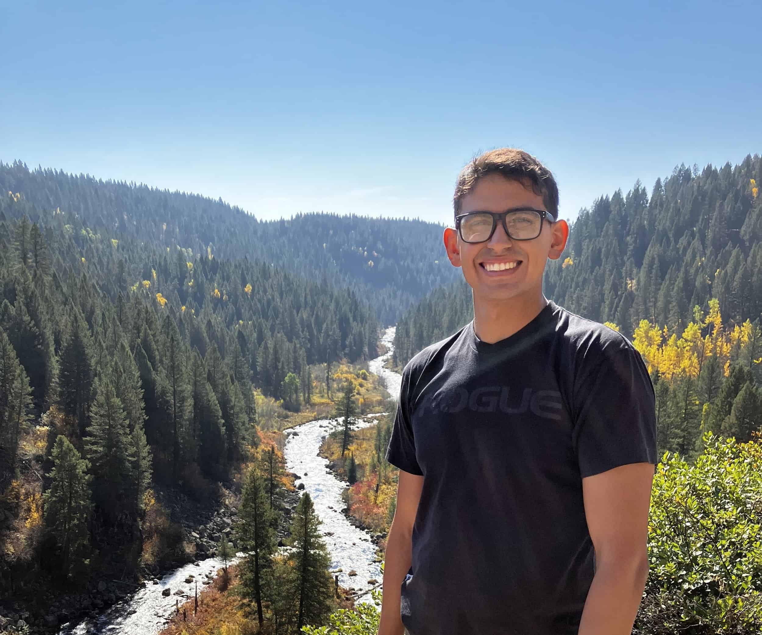 Young man smiling in front of mountain river and forest landscape.