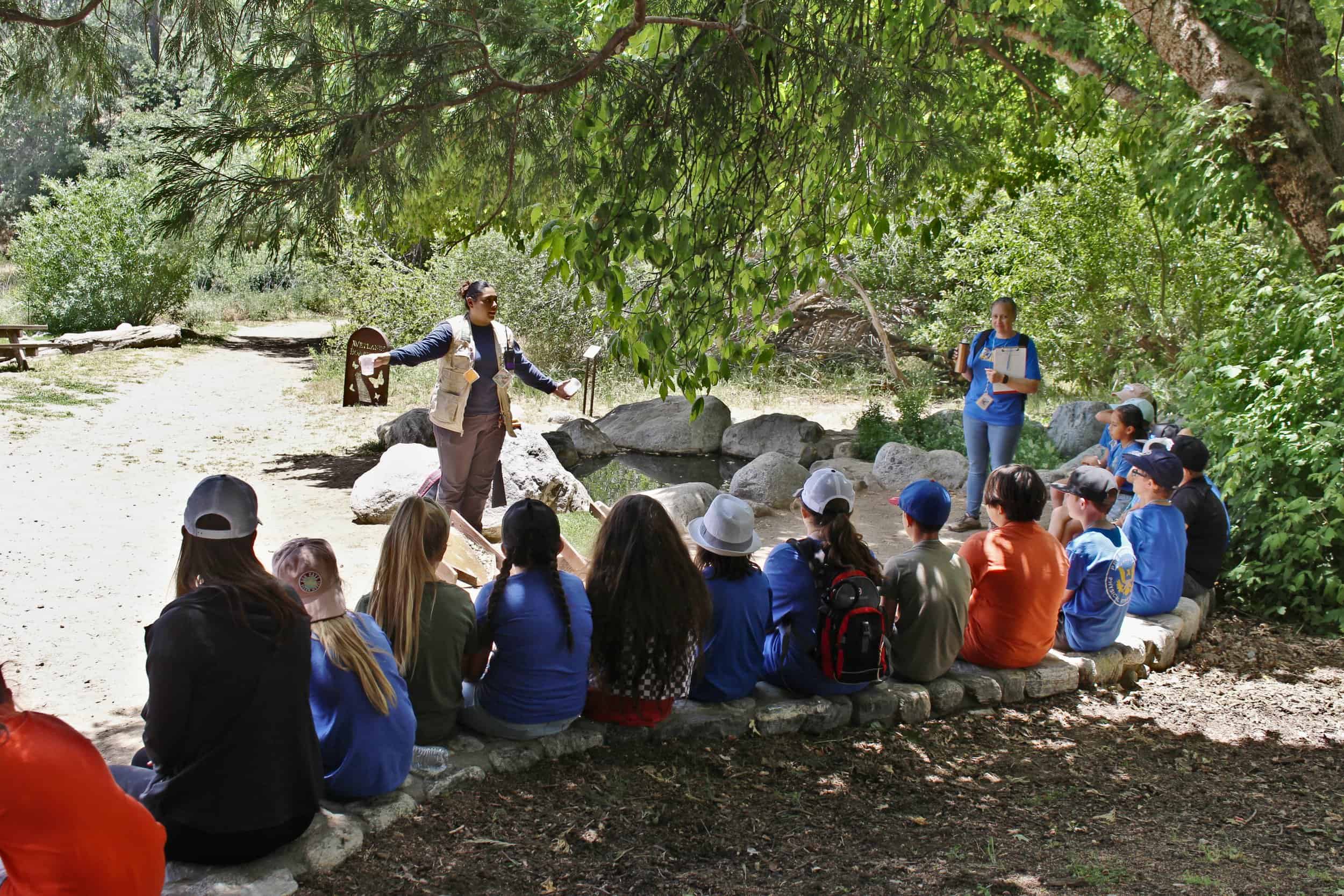 Group of children and adults participating in outdoor nature education under a tree.