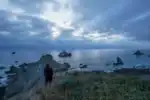 Person overlooking rocky coastline during cloudy sunset.