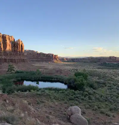 Desert landscape with cliffs, small pond, and open sky at sunset.