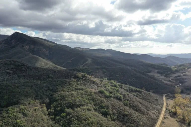 A lush mountain landscape with rolling hills and a winding dirt trail under a partly cloudy sky.