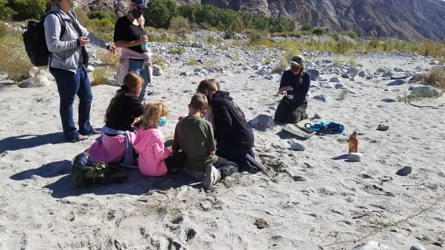 Group of children and adults participating in outdoor educational activity on a rocky riverbank.