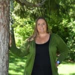 Woman in green cardigan standing beside a tree in a lush forest setting.