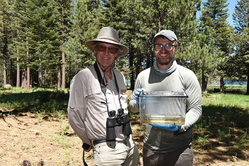 Two men in outdoor gear holding a fish tank in a forest setting.