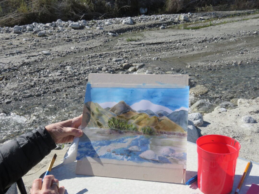 Watercolor painting of mountains and river on outdoor canvas with art supplies nearby.