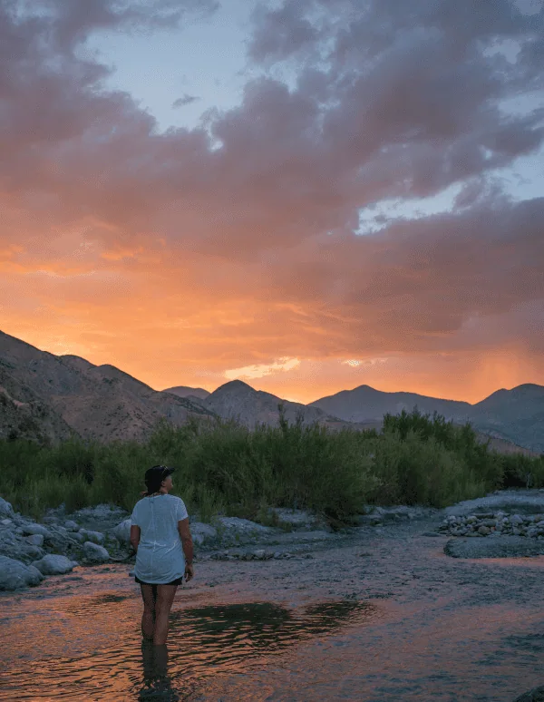 Person standing in river at sunset with mountains in background.